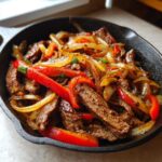 Close-up of seasoned steak fajitas with caramelized onions and bright red bell peppers served in a black cast iron skillet.