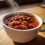 A steaming white mug filled with rich, hearty chili featuring ground meat and beans, showcasing the best chili recipe.