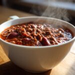 Close-up of a steaming white bowl filled with rich, hearty chili, featuring ground meat and kidney beans.