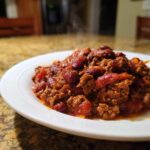 Close-up of a hearty serving of steaming crockpot chili loaded with ground beef and kidney beans on a white plate.