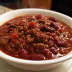 Close-up of a steaming white bowl filled with rich, thick crockpot chili featuring ground beef and red kidney beans.