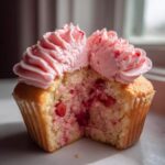 Close-up of a strawberry cupcake cut in half showing moist cake studded with strawberry pieces and topped with pink strawberry frosting.