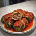 A plate of homemade cabbage rolls covered in rich tomato sauce, one cut open showing the savory ground meat filling.