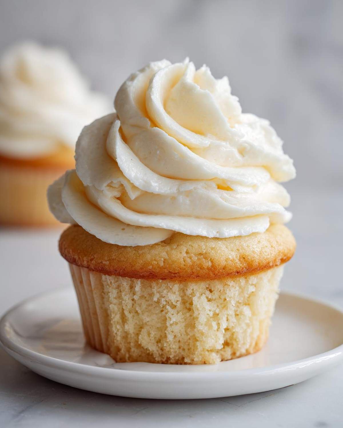 A close-up of a moist vanilla cupcake topped with a generous swirl of creamy white vanilla frosting.
