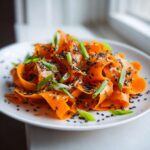 Close-up of a vibrant raw carrot salad made with shaved carrot ribbons, topped with black sesame seeds and chopped green onions.