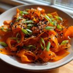 Close-up of a vibrant raw carrot salad featuring shaved carrot ribbons topped with black and white sesame seeds and sliced green onions.