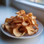 A pile of crispy, curled Air Fryer Cinnamon Sugar Apple Chips dusted heavily with cinnamon sugar on a light plate.