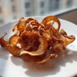 A pile of crispy, curled Air Fryer Cinnamon Sugar Apple Chips dusted with spice on a white plate.
