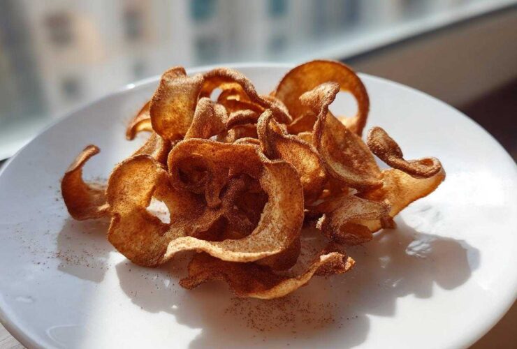 A pile of crispy, curled Air Fryer Cinnamon Sugar Apple Chips dusted with spice on a white plate.