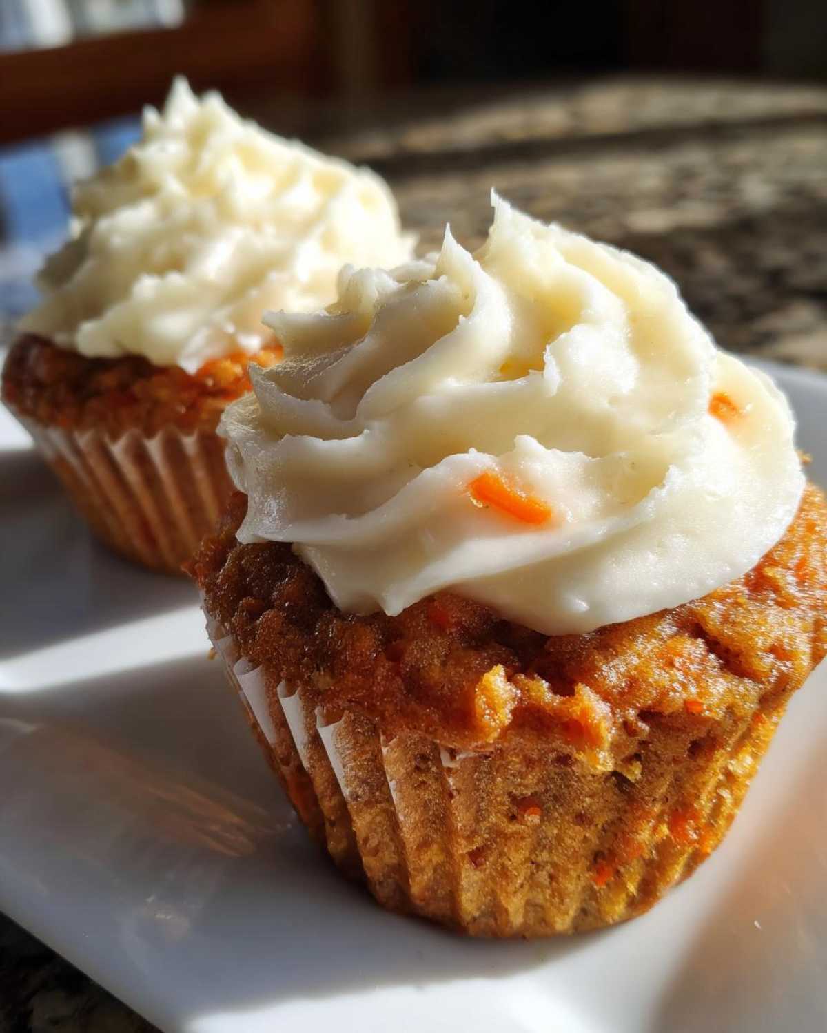 Two moist Gluten-Free Carrot Cake Cupcakes topped with thick swirls of cream cheese frosting, sitting on a white plate.