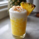 A tall glass featuring an Iced Pineapple Coconut Mocktail, topped with white foam and toasted coconut flakes.