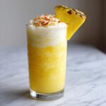 A tall glass containing a vibrant Iced Pineapple Coconut Mocktail, topped with foam and toasted coconut.