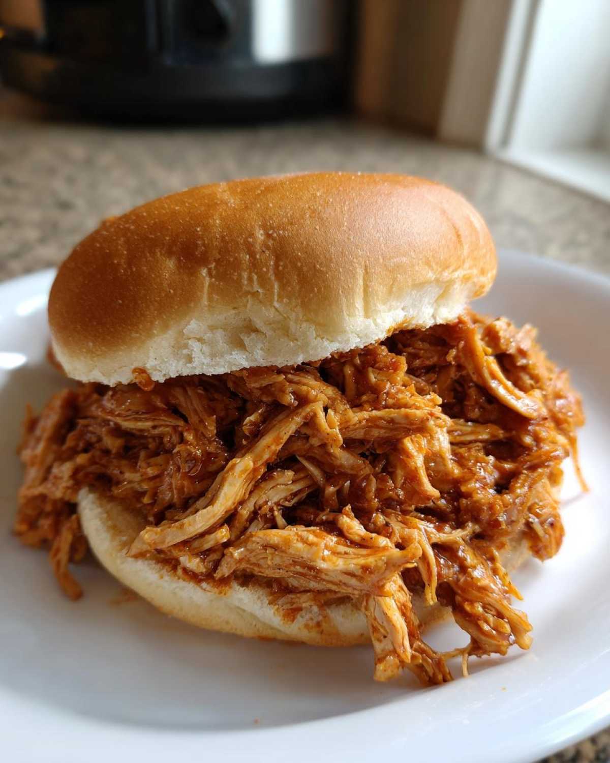 Close-up of a delicious Instant Pot BBQ Pulled Chicken sandwich piled high on a soft white bun, served on a white plate.