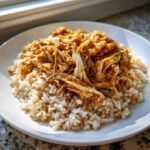 Close-up of shredded, saucy chicken piled high on a bed of brown rice in a white bowl.