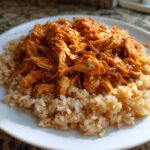 A white plate featuring a bed of brown rice topped with saucy, shredded chicken, ready for Instant Pot Chicken Burrito Bowls.