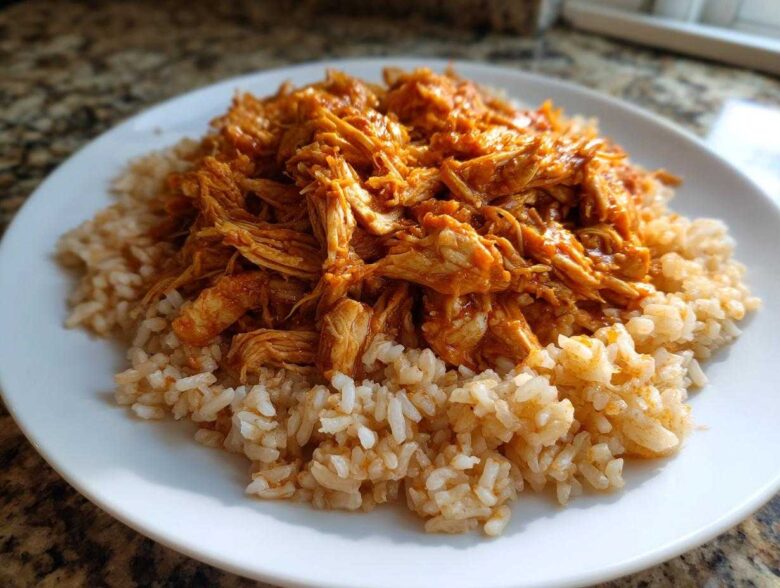 A white plate featuring a bed of brown rice topped with saucy, shredded chicken, ready for Instant Pot Chicken Burrito Bowls.