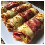 Four baked Keto Italian Sub Roll-Ups wrapped in salami and melted cheese, sprinkled with herbs.
