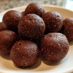 A close-up of several rich, dark brown No-Bake Brownie Batter Protein Balls stacked on a white plate.