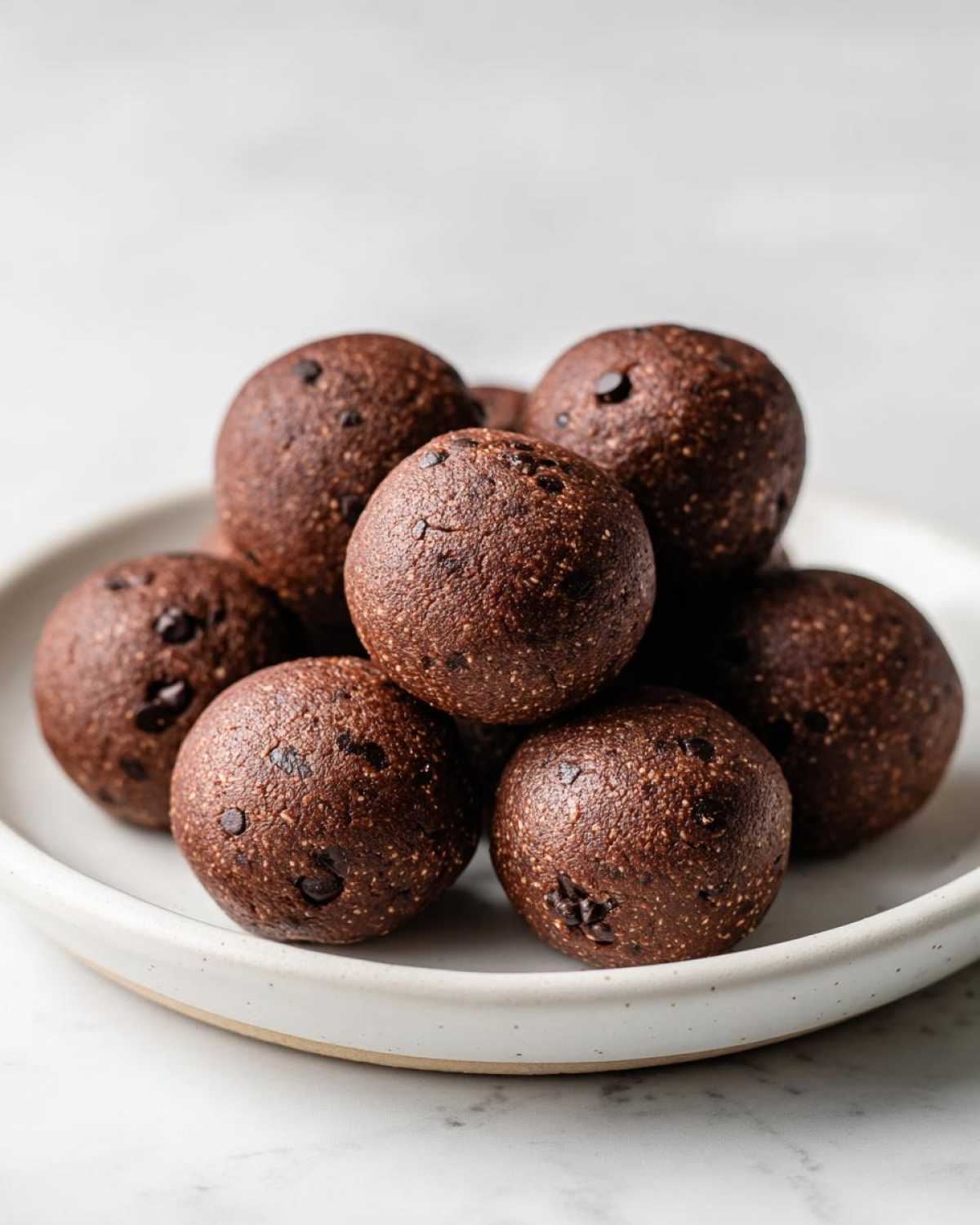 A close-up of several rich, dark brown No-Bake Brownie Batter Protein Balls piled on a light, speckled ceramic plate.