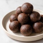A stack of rich, dark chocolate No-Bake Brownie Batter Protein Balls piled on a light ceramic plate.