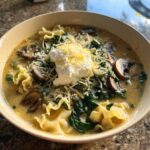 A close-up of One-Pot Creamy Spinach Mushroom Lasagna Soup topped with ricotta and shredded cheese.