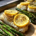 Close-up of flaky Sheet Pan Lemon Herb Cod with Asparagus topped with lemon slices and herbs.
