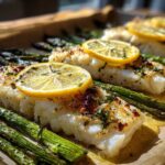 Close-up of flaky Sheet Pan Lemon Herb Cod fillets topped with lemon slices and served alongside roasted asparagus.