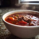 Close-up of a steaming white bowl filled with rich, red Keto Taco Soup with Ground Beef and visible chunks of vegetables.