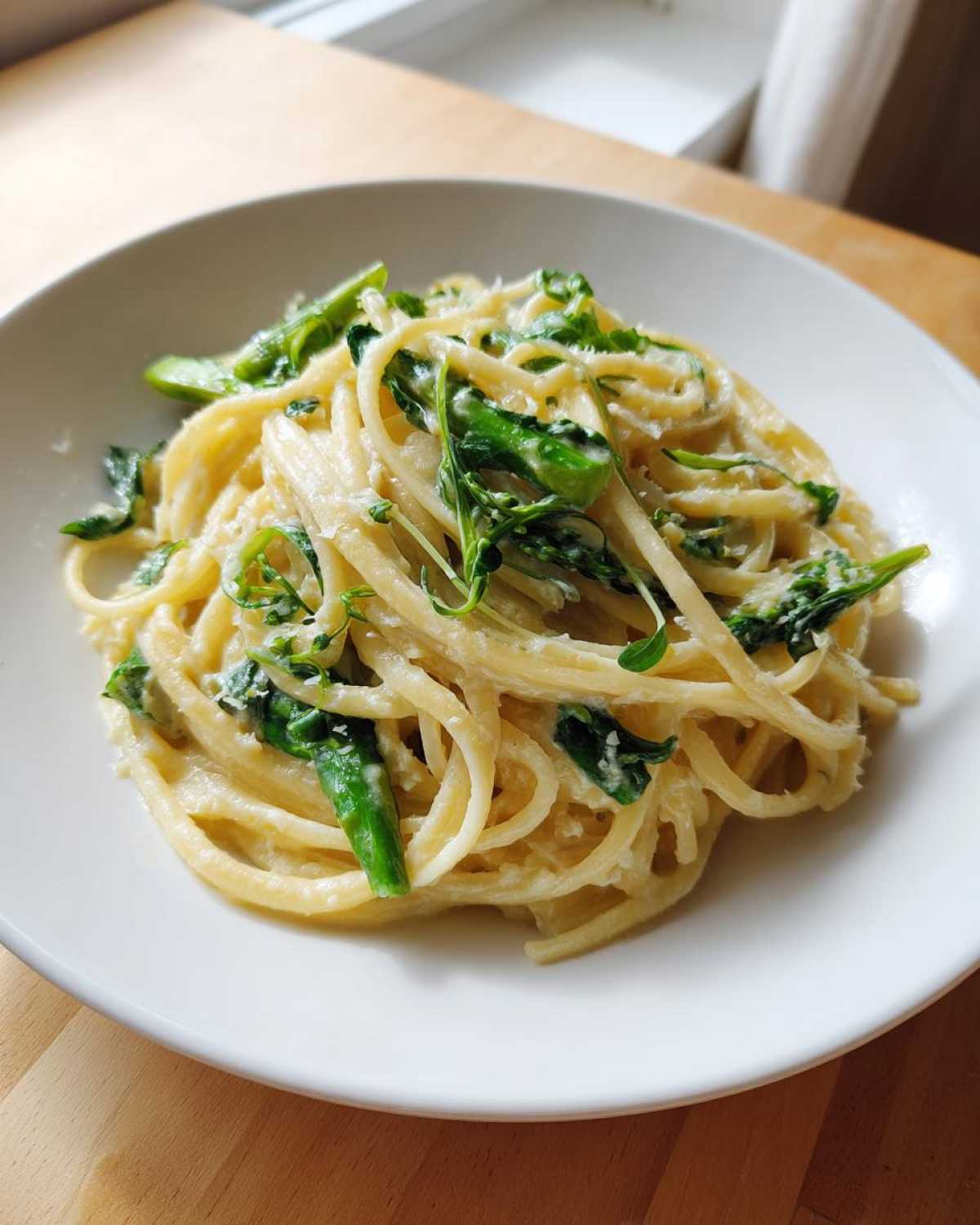 A close-up of creamy Vegan Lemon Asparagus Pasta with bright green asparagus pieces and pea shoots on a white plate.