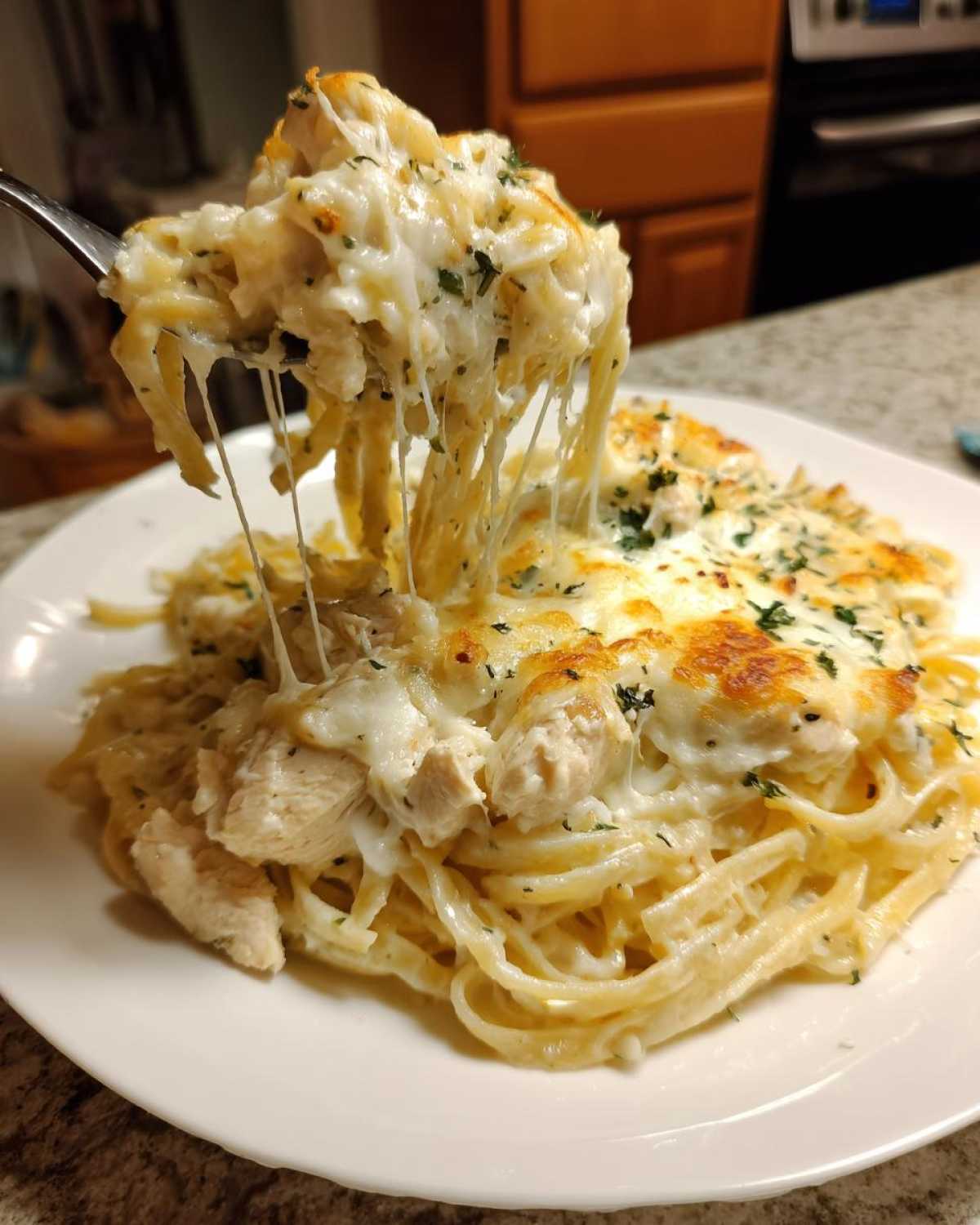A fork lifts a serving of Cheesy Chicken Alfredo Bake with Mozzarella, showing long, dramatic cheese pulls over pasta and chicken chunks.
