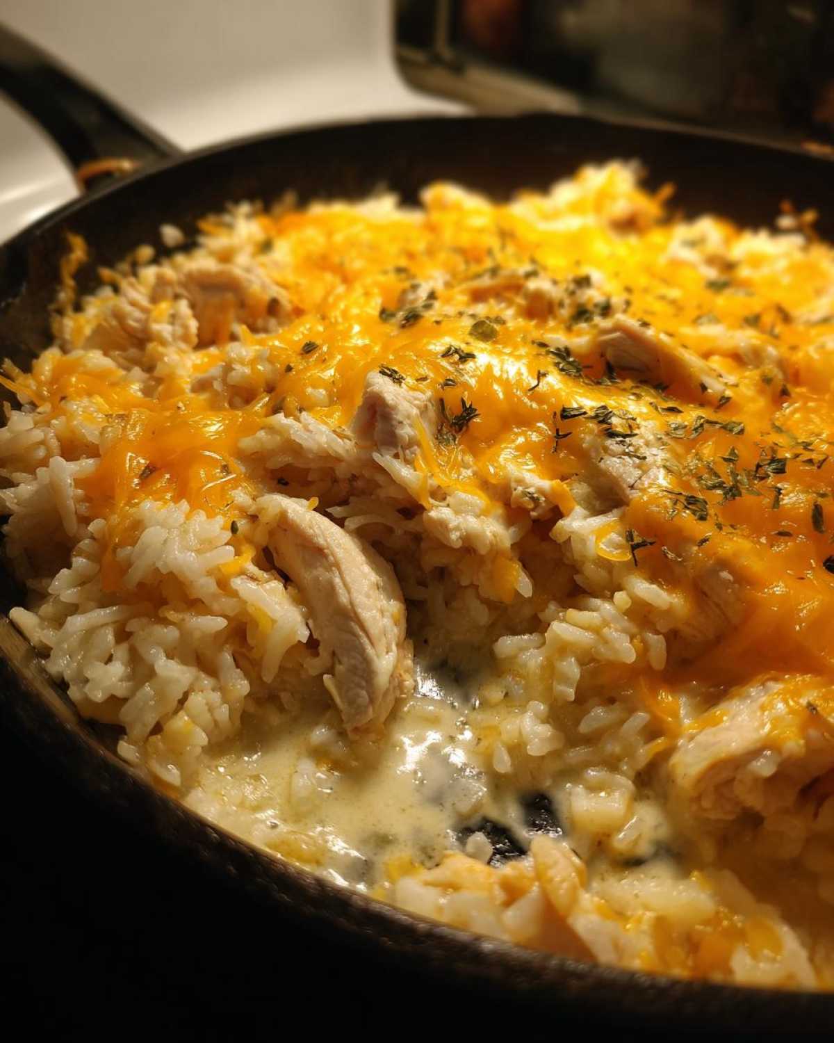 Close-up of Cheesy Chicken and Rice Skillet in a dark pan, topped with melted orange herbs.