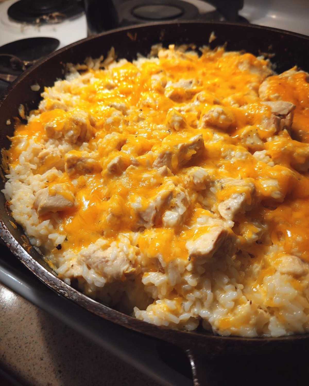 Close-up of Cheesy Chicken and Rice Skillet topped with melted orange cheddar cheese in a dark pan.