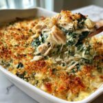 A wooden spoon lifts a scoop of creamy Cheesy Chicken and Spinach Bake Comfort Meal, showing shredded chicken and melted cheese strings.