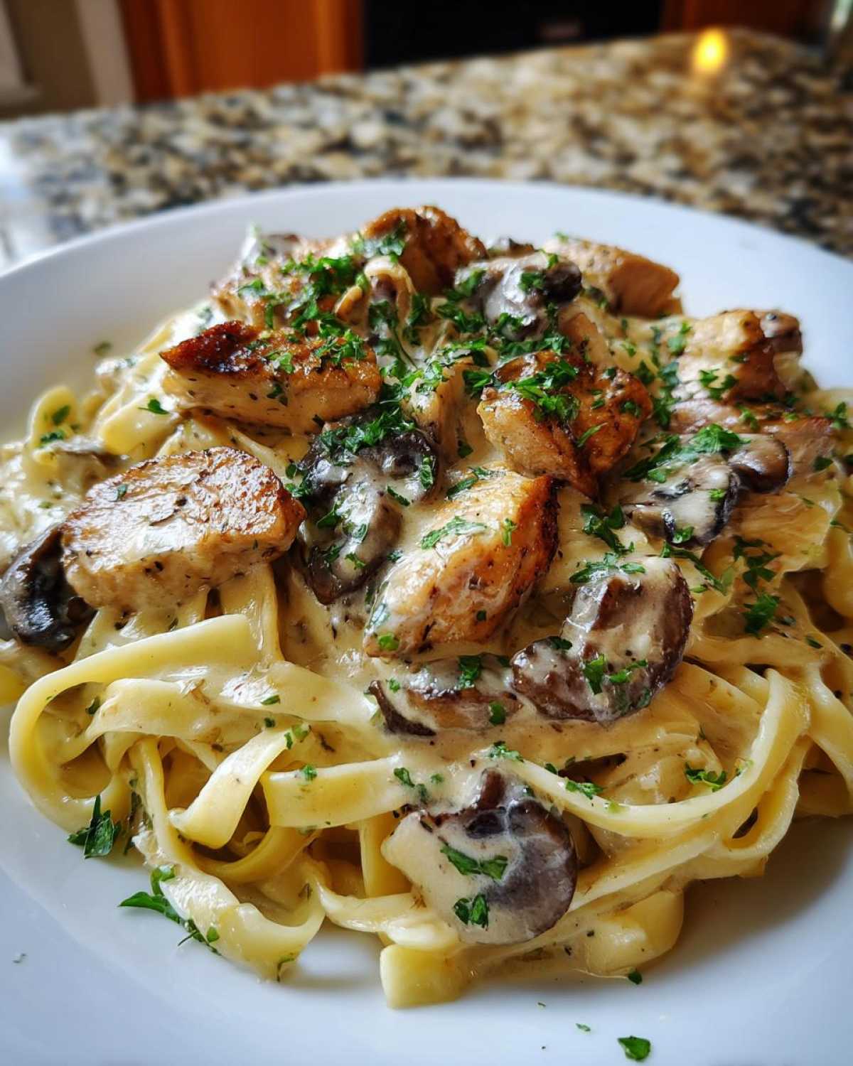 A close-up of a white plate filled with fettuccine pasta coated in a creamy garlic sauce, topped with seared chicken pieces and mushrooms, garnished with parsley.