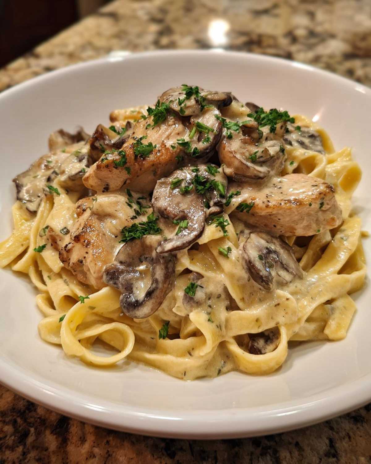 A close-up of fettuccine pasta topped with seared chicken pieces, sautéed mushrooms, and rich garlic sauce, garnished with parsley for Creamy Mushroom Chicken Pasta.