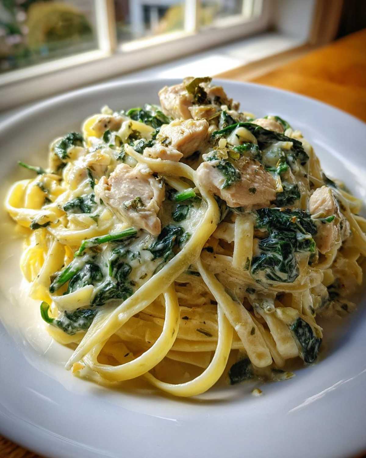 A close-up of creamy spinach pasta with chicken dinner served on a white plate near a window.