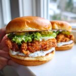 A hand holding a delicious Crispy Chicken Burger with Garlic Mayo, lettuce, and pickles on a brioche bun.