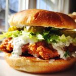 A close-up of a Crispy Chicken Burger with Garlic Mayo, featuring a golden fried patty, lettuce, pickles, and creamy sauce on a toasted bun.