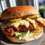 A close-up of a perfectly assembled Crispy Chicken Burger with Honey Mustard Sauce dripping over the fried patty and pickles.