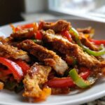 Close-up of crispy chicken fajitas strips mixed with sautéed red, green, and orange peppers on a white plate.