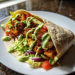 A close-up of a Crispy Chicken Wrap with Avocado Sauce drizzled over seasoned chicken, lettuce, tomato, and red onion.