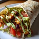 A close-up of a perfectly assembled Crispy Chicken Wrap with Avocado Sauce, filled with seasoned chicken, lettuce, tomato, and onion.