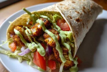 A close-up of a perfectly assembled Crispy Chicken Wrap with Avocado Sauce, filled with seasoned chicken, lettuce, tomato, and onion.