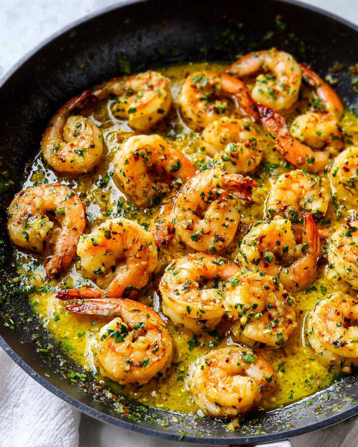Close-up of plump shrimp simmering in a rich, yellow garlic butter sauce seasoned with fresh parsley in a black skillet.