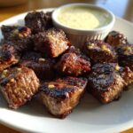 Close-up of juicy Garlic Butter Steak Bites seasoned with minced garlic, served on a white plate with a side of creamy dipping sauce.