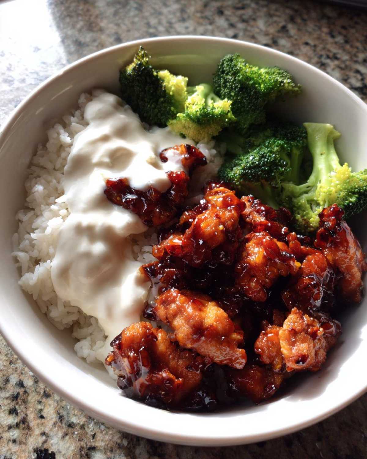A close-up of a Honey Garlic Chicken Bowl with rice, steamed broccoli, and a dollop of creamy white sauce.