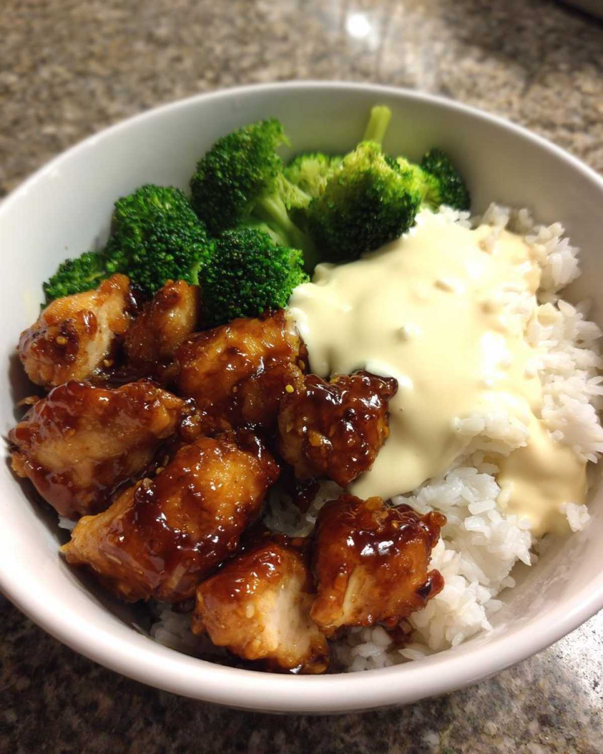 A close-up of a Honey Garlic Chicken Bowl featuring glazed chicken pieces, white rice, steamed broccoli, and a drizzle of creamy sauce.