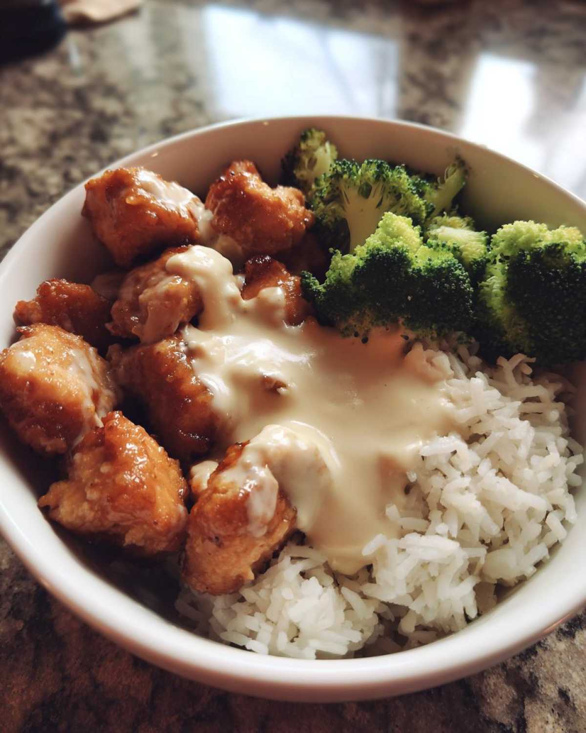 A close-up of a Honey Garlic Chicken Bowl with crispy chicken pieces, white rice, steamed broccoli, and a generous drizzle of creamy sauce.