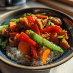 A close-up of a Honey Garlic Chicken Bowl featuring crispy chicken, red peppers, carrots, and snow peas over white rice.