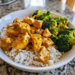 A close-up of a Honey Mustard Chicken Bowl with Rice, featuring chunks of chicken coated in yellow sauce next to bright green broccoli florets.